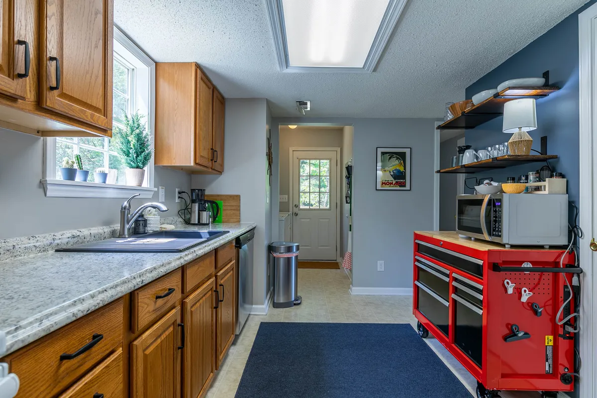 A functional kitchen countertop is the focal point, with a red tool cart, plants in the window, and a doorway leading to the outdoors. The scene blends functionality with homely charm, suggesting a place for baking, meal prep, and quick transitions between indoor and outdoor cooking.