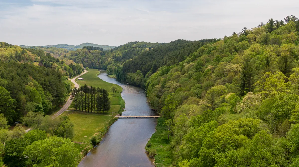 A serene river winds past a bridge and through the hills, with lush trees lining the banks. The natural beauty of the area is on full display, suggesting opportunities for relaxation and outdoor activity.