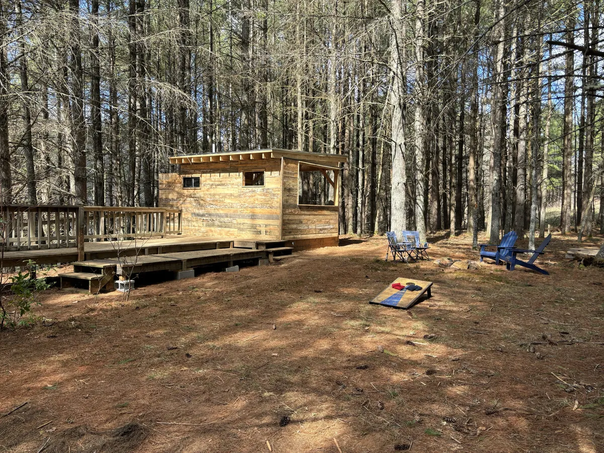 A wooden cabin decked with Adirondack chairs and a cornhole game, set against a peaceful backdrop of trees. The deck area is perfect for relaxing, playing outdoor games, and sharing time with friends and family in a serene environment.