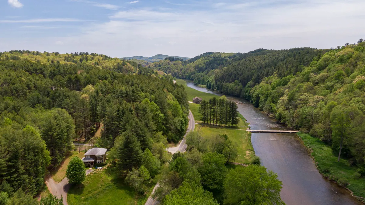 The image showcases a tranquil scene with the New River winding through dense forests and scenic hills, as the sun sets over the water. The cozy cabin sits nestled amidst three private acres, offering sweeping views of the river and surrounding wilderness. Outdoor activities like kayaking, hiking, and fishing are readily accessible, and wildlife can be seen in the fading light.