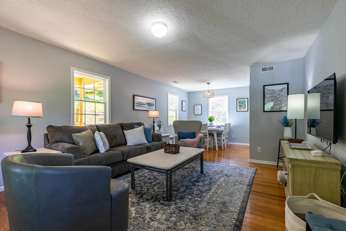 A welcoming open-plan living room featuring a plush sofa, a coffee table, and a view toward a cozy dining area. Large windows flood the space with natural light, highlighting the warm, inviting atmosphere ideal for relaxations and gatherings.