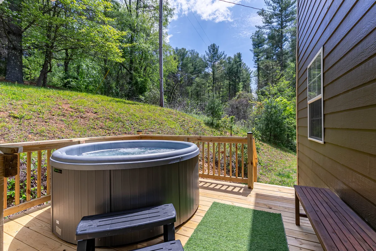 A hot tub sits on a wooden deck, framed by lush green grass and tall trees, with ample space around for socializing or simply taking in the views. The tranquil setting is perfect for relaxation.