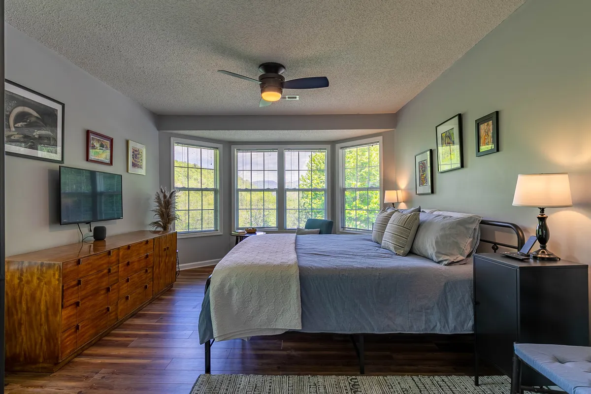 This image shows a bedroom with a cozy bed positioned near generous bay windows, a dresser, tasteful artwork on the walls, and a nightstand with a lamp. Sunlight pours into the room, creating a serene and restful ambiance designed for relaxation or quiet activities like reading or watching television.
