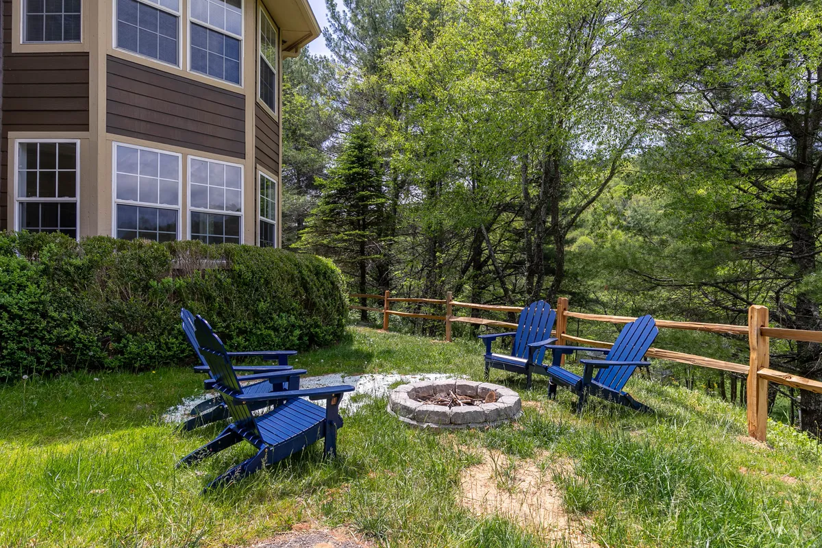 Several blue Adirondack chairs encircle a fire pit set on green grass, with the house and surrounding trees in the background. The area is clearly designed for communal gatherings and outdoor storytelling under the stars.