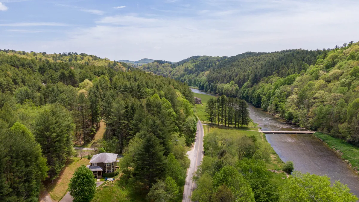 A sweeping view of the river, wooded hills, and the house nestled within the landscape. The composition emphasizes the peacefulness and beauty of the location, showing river vistas and expansive natural surroundings that invite outdoor exploration and morning coffee on the porch.