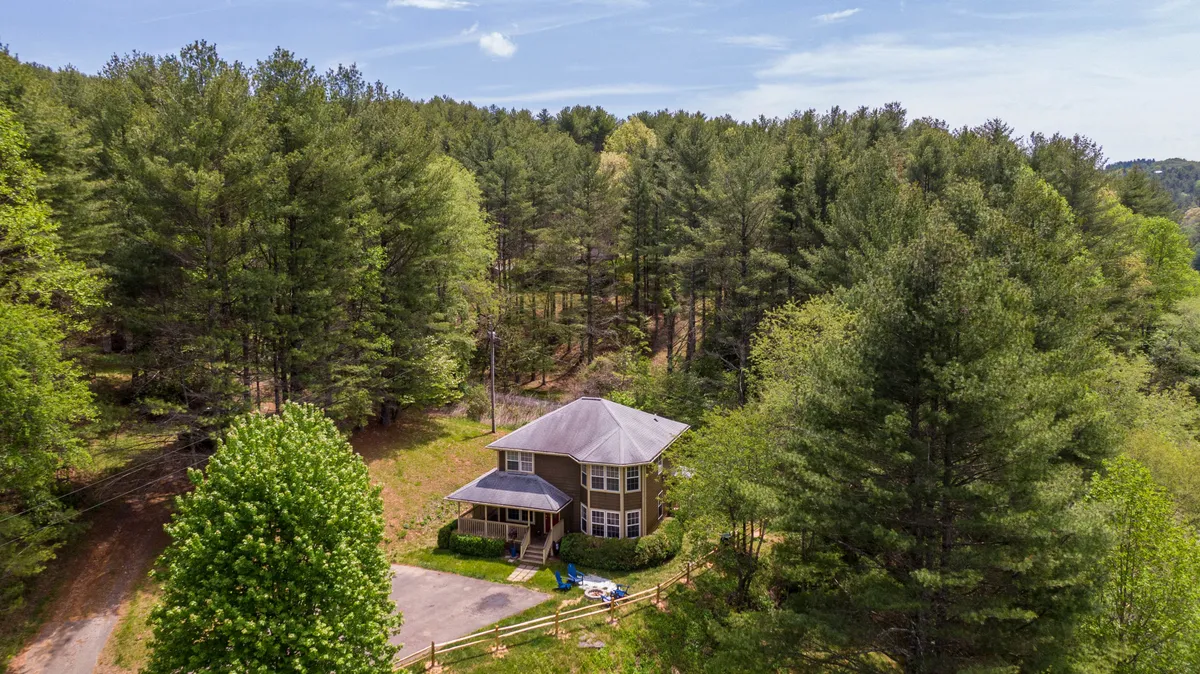 An inviting exterior shot showcasing the house surrounded by tall trees, with a glimpse of the driveway and the spacious porch. The setting feels secluded yet welcoming, perfect for nature walks, outdoor gatherings, and enjoying the quiet ambiance.