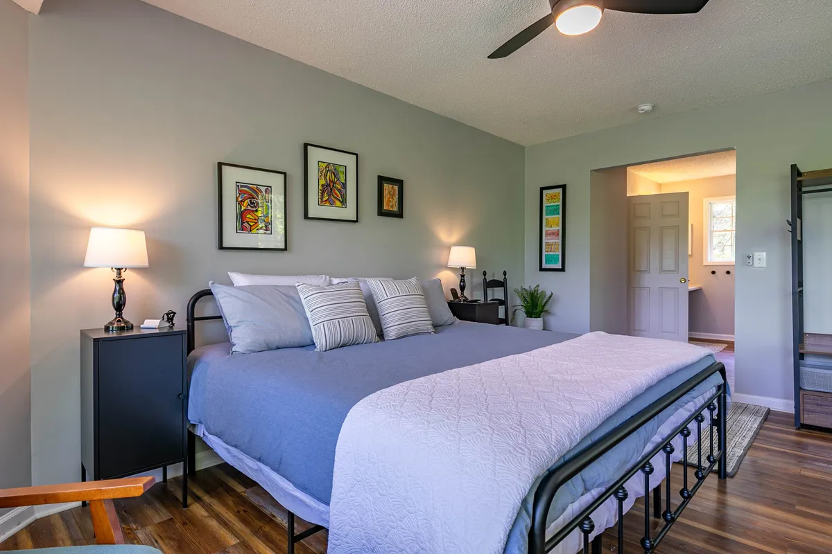 A comfortable bedroom scene featuring a well-made bed with decorative bedding, artwork on the walls, and matching nightstands topped with lamps. The space feels cozy, inviting, and ideal for unwinding.
