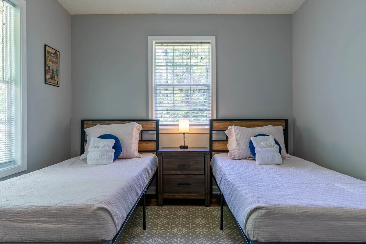 A bright, airy bedroom with two twin beds, a nightstand with a lamp between them, and a window letting in natural light. The arrangement invites both rest and conversation, making it ideal for children or friends sharing the space.