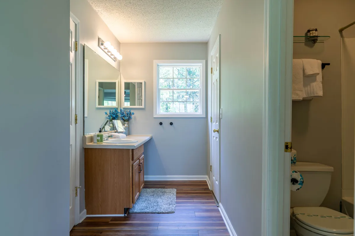 A bathroom with a vanity, sink, and mirror, positioned near a window that lets in natural light and decorated with fresh flowers. The overall mood is serene and inviting, suggesting a pleasant place for morning or evening routines.