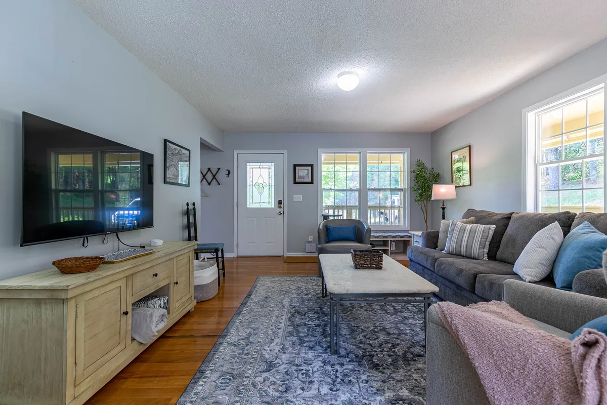 A cozy living area furnished with a comfortable sofa, coffee table, and a Smart TV positioned near windows and the front door. The space feels intimate yet open, with plenty of natural light and areas for both relaxation and entertainment.