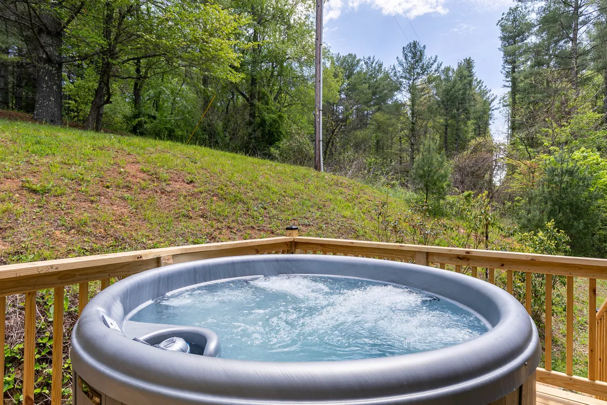 A tranquil exterior scene featuring a wooden deck with a 5-person Nordic hot tub surrounded by lush greenery. The deck is set against a backdrop of trees, evoking a sense of privacy and immersion in nature. The hot tub is the focal point, suggesting relaxation and stargazing opportunities.