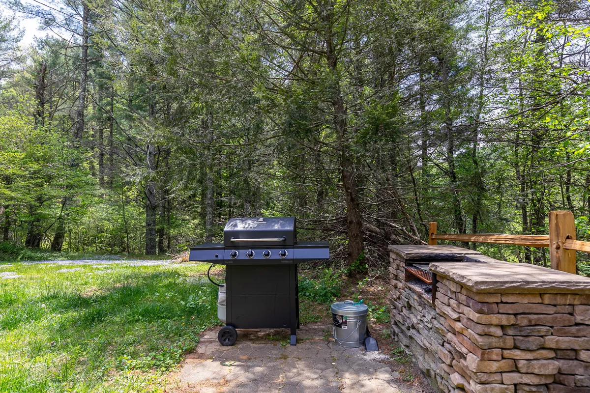 An exterior shot featuring a sturdy grill set against a rustic stone structure, surrounded by trees. The scene suggests gatherings centered around barbecuing and enjoying the fresh mountain air.