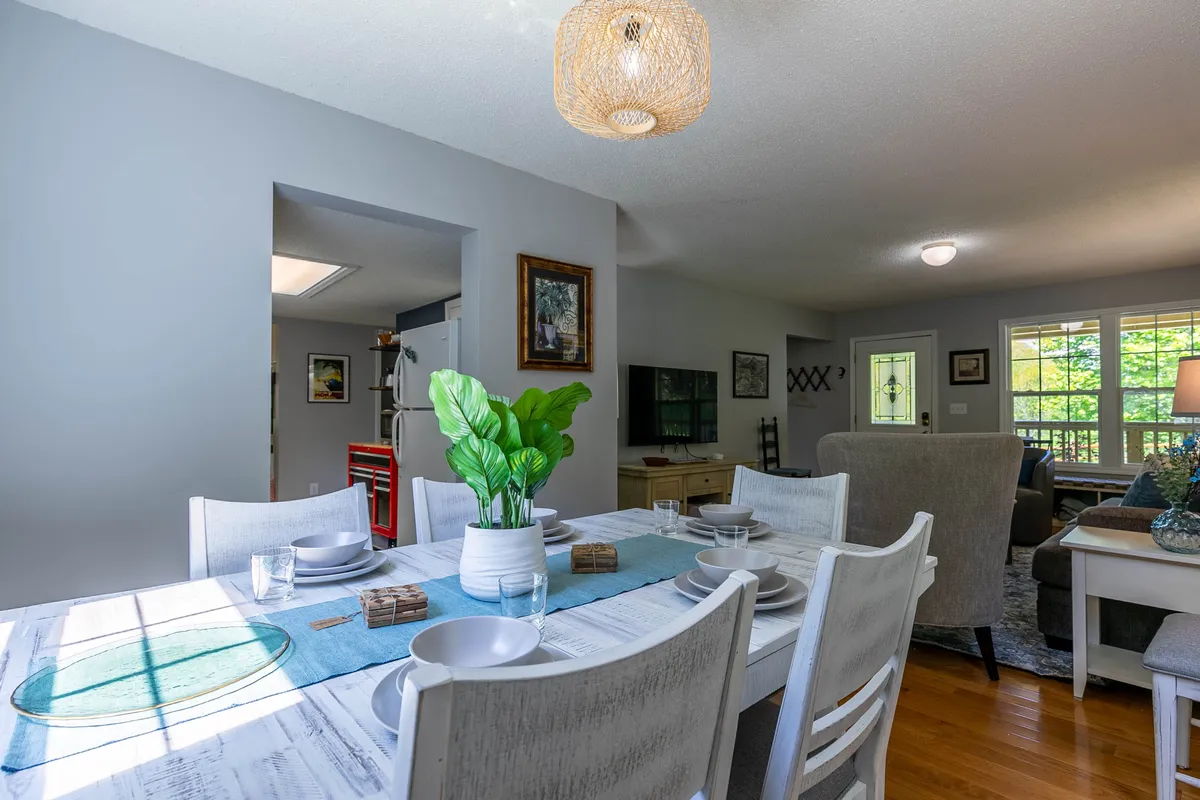 The focus is on a welcoming dining table set with a green plant centerpiece, complemented by a stylish light fixture above, artwork on the wall, and a sofa in the background. The space exudes warmth and is ideal for family meals, gatherings, and leisurely brunches.